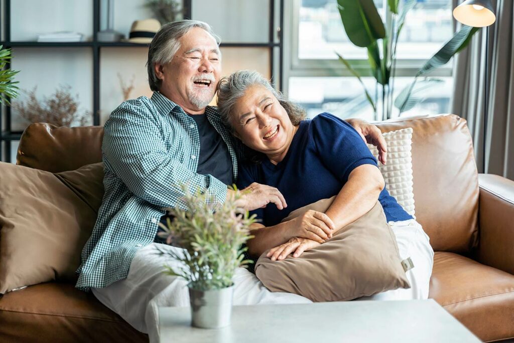 Sage Oak of Denton | Happy senior couple smiling and laughing together on couch in living room